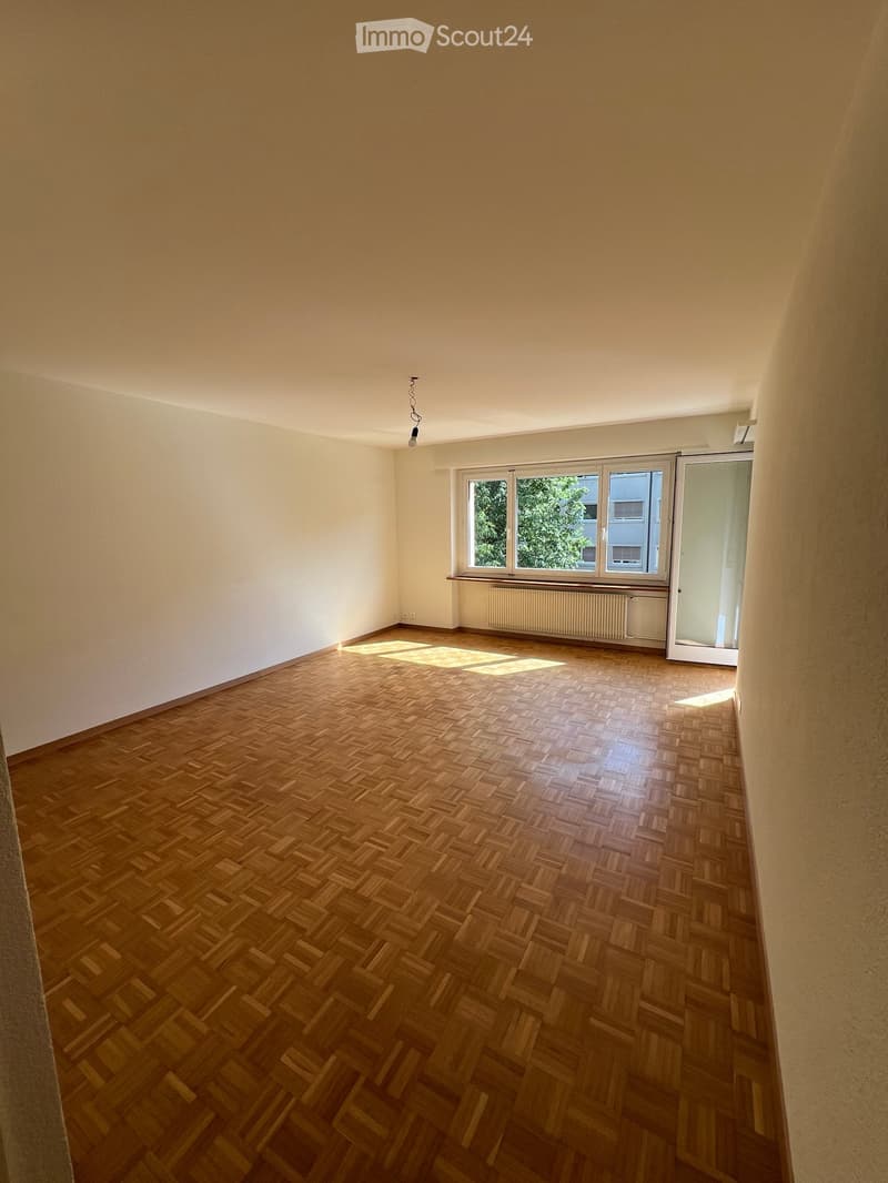 Empty room with wooden herringbone flooring, white walls, large window with radiator, and a sliding glass door leading outside.