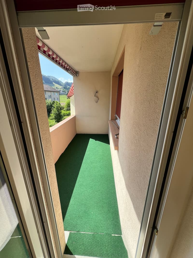 Balcony with green artificial turf flooring, beige walls, and a view of a landscape with trees and houses.