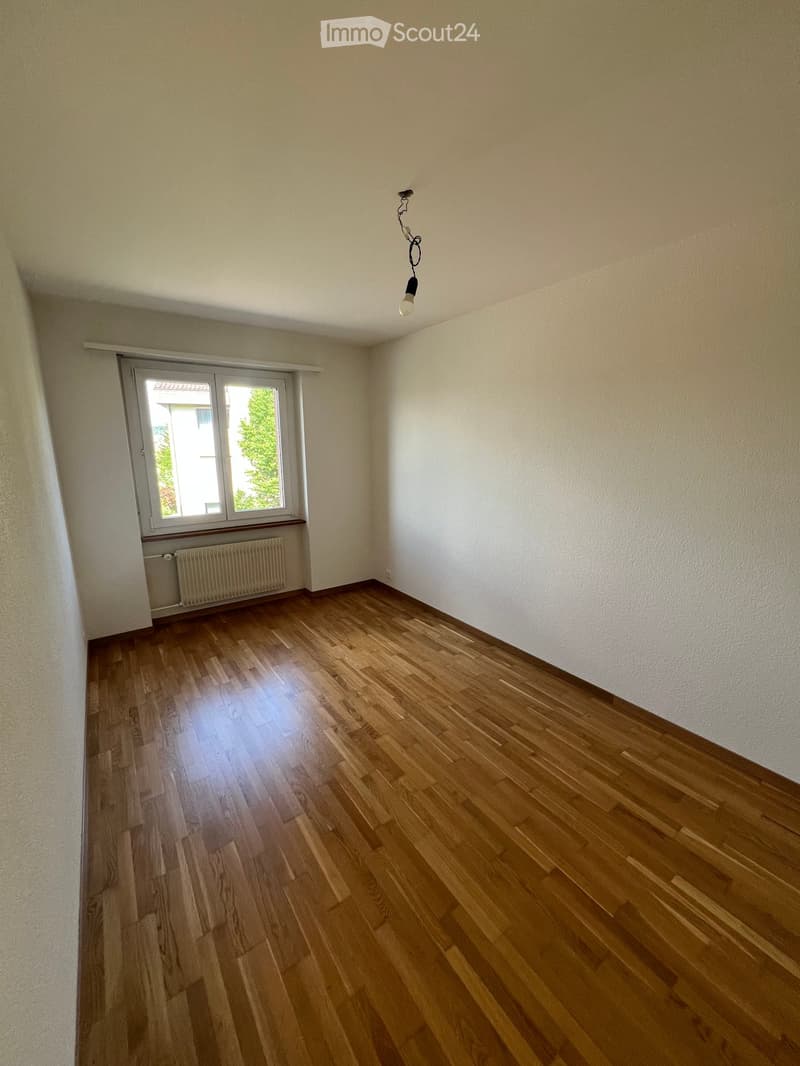 Empty room with wooden flooring, white walls, a window with a radiator below, and a single hanging light fixture.