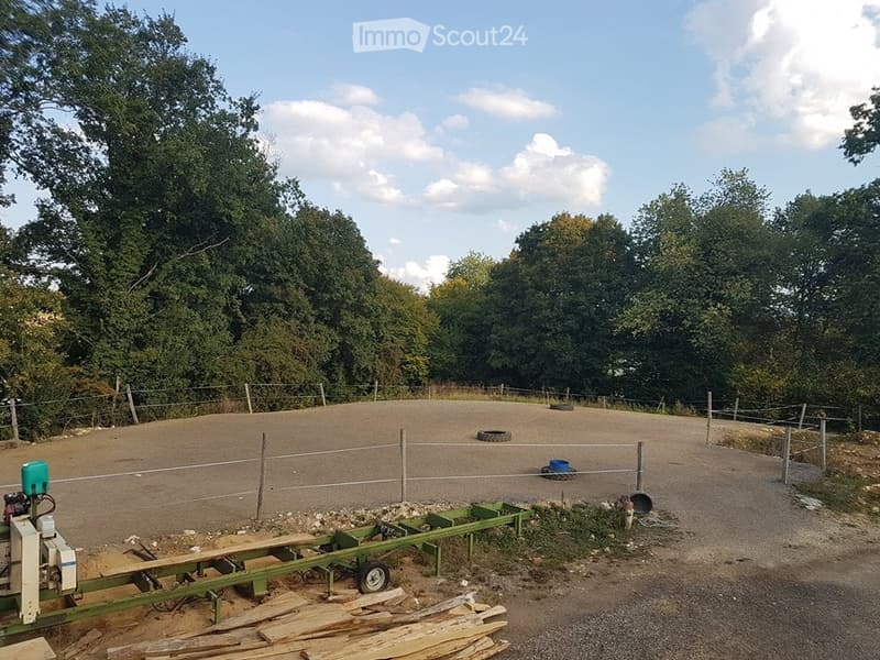 Outdoor area enclosed by a wire fence, with a dirt ground, scattered tires, and a small green trailer with equipment.