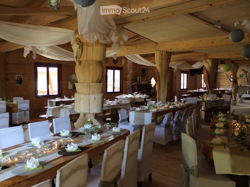 A spacious, rustic dining area with wooden tables and chairs, set for a formal event. The room features exposed wooden beams, large windows, and decorative elements like hanging hearts and a central wooden pillar.