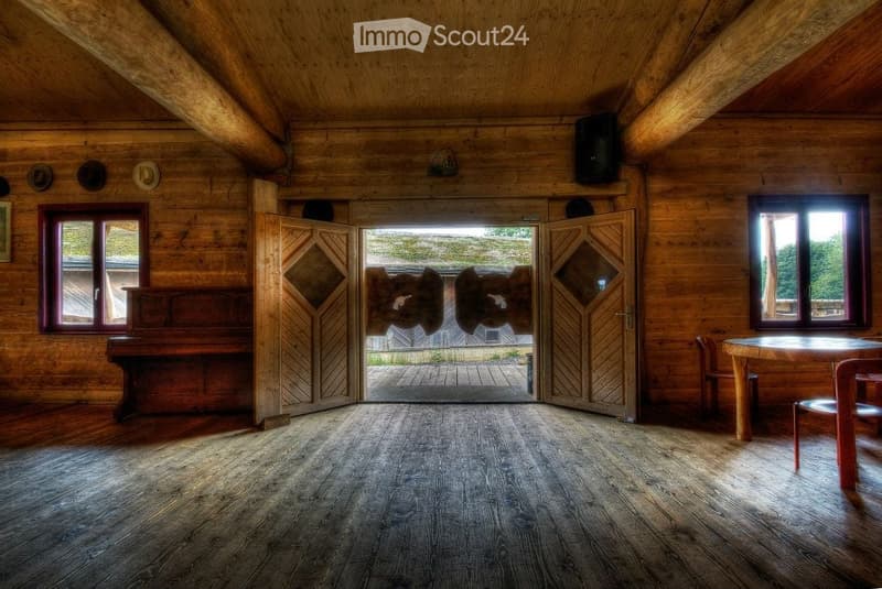 Large, open wooden room with exposed beams, wooden floor, large double doors leading outside, and a few windows. The room is completely empty with no furniture.