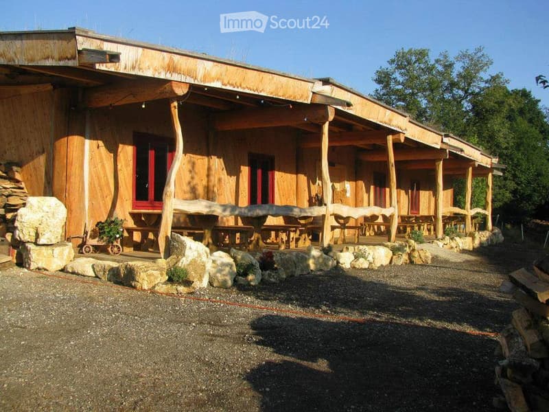 A rustic wooden building with a covered porch area, featuring wooden benches and tables, surrounded by large stones and greenery.