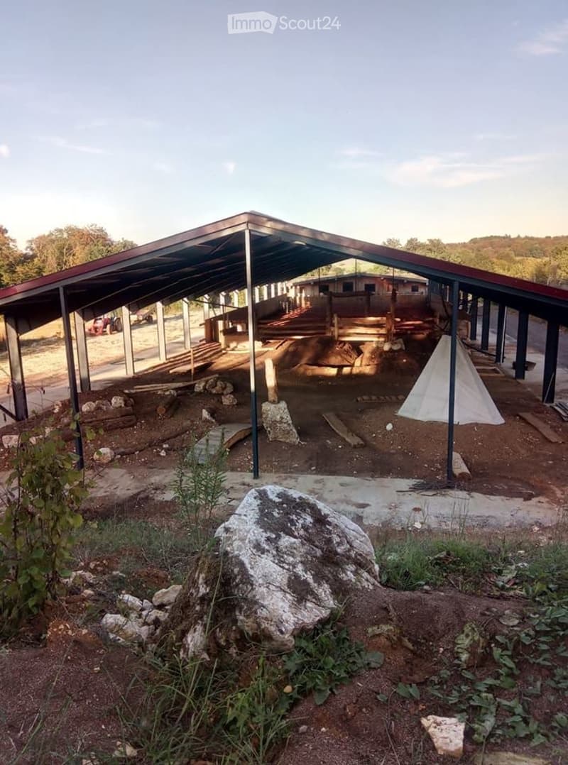 An outdoor area with a metal canopy structure, dirt ground, scattered rocks, and a small white tent. There are wooden planks and a few vehicles in the background.