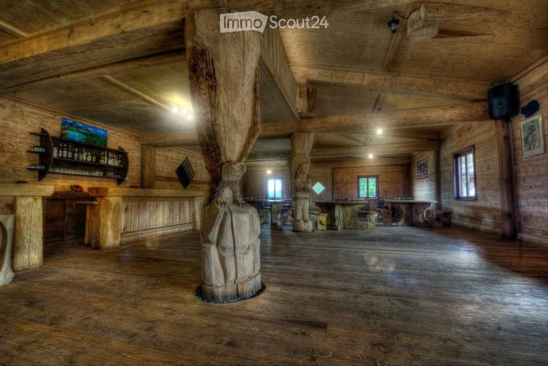A spacious wooden interior with exposed beams and large wooden pillars, featuring a bar area with shelves, a central table, and chairs, illuminated by warm lighting.
