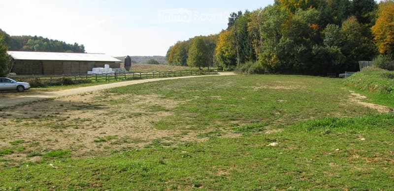 Open grassy field with a dirt path, surrounded by trees and a wooden fence. A barn and a parked car are visible in the background.