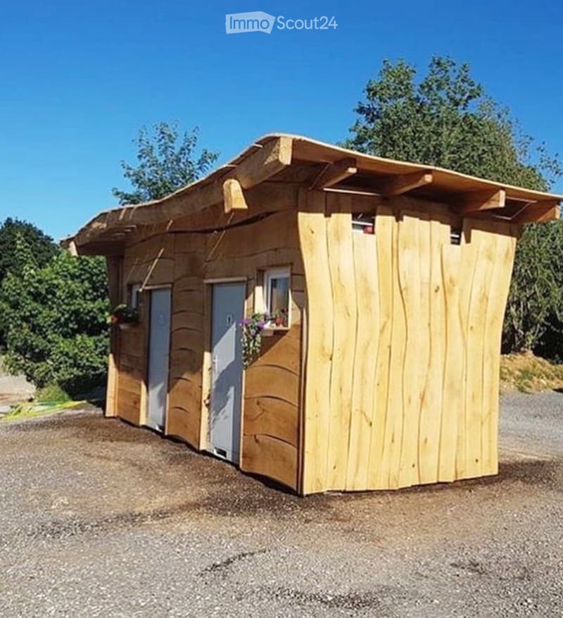 A small wooden cabin with a sloped roof, two doors, and a window with a flower box, situated on a gravel surface with trees in the background.
