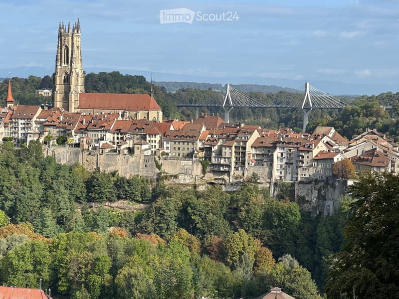 A panoramic view of a historic town with red-roofed buildings, a prominent church tower, and a modern suspension bridge spanning a river valley.