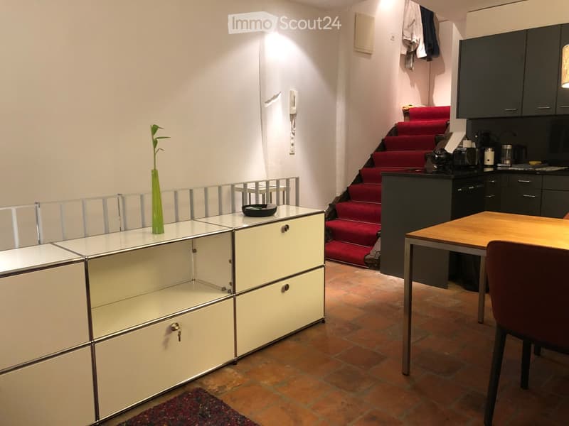 A modern, well-lit room with a wooden table, a chair, a white cabinet with glass doors, a green vase on the cabinet, and a staircase with red carpeting leading upwards.