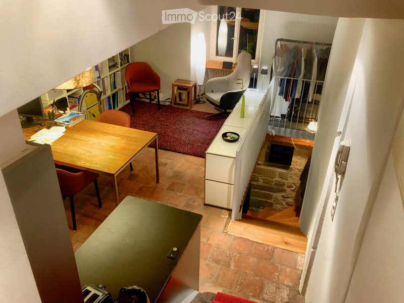 A multi-functional room with a wooden table, red chairs, a sofa, a lamp, a bookshelf, and a small cabinet. The space features a mix of tiled and wooden flooring.
