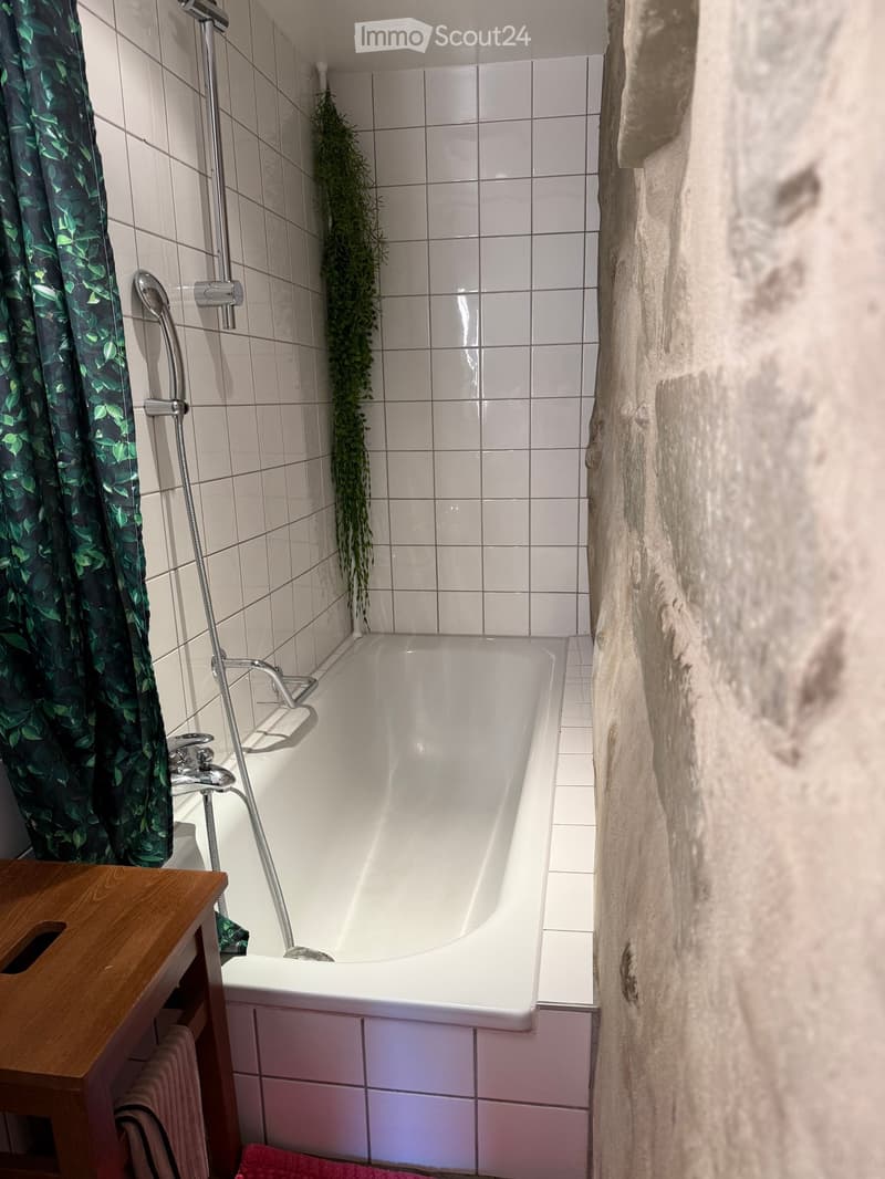 Bathroom with a white bathtub, tiled walls, a wooden shelf with towels, and a green leafy shower curtain.