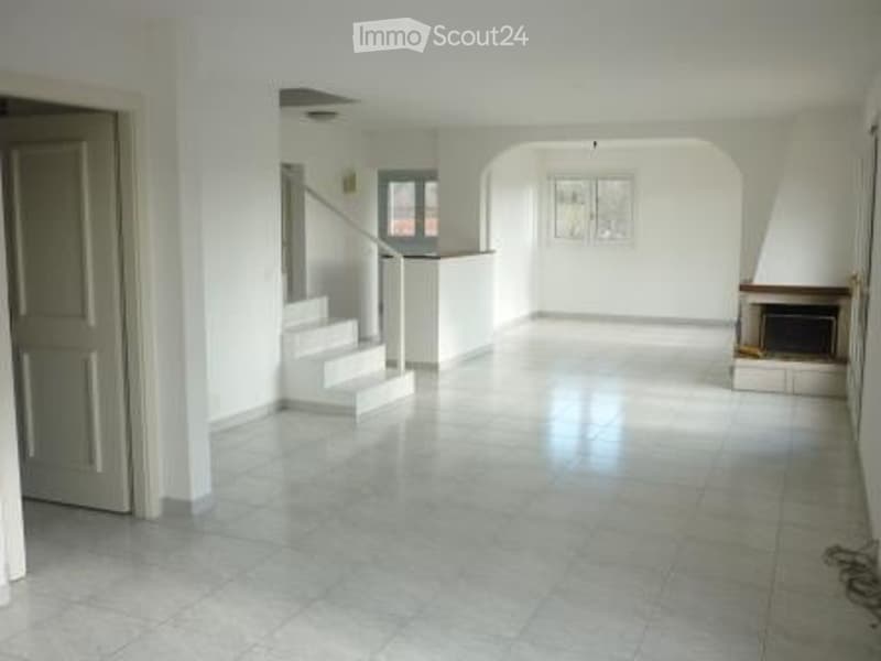 Empty living space with white walls, tiled floor, staircase leading to an upper level, fireplace, and large windows.