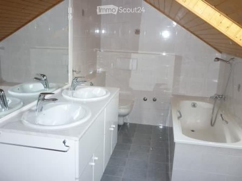 Modern bathroom with two sinks, a bathtub, and a toilet. The room features white tiles and a wooden slanted ceiling.