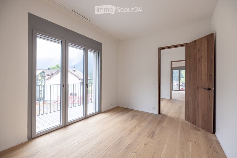 Empty room with light wooden flooring, white walls, large sliding glass doors leading to a balcony, and an open wooden door to another room.