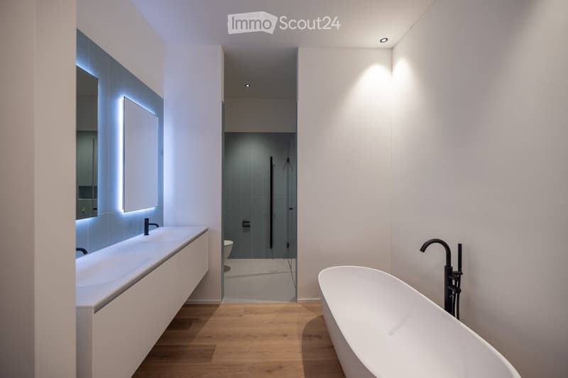Modern bathroom with a freestanding bathtub, a sleek sink with a modern faucet, and a large mirror with integrated lighting. The space features light wooden flooring and recessed ceiling lighting.