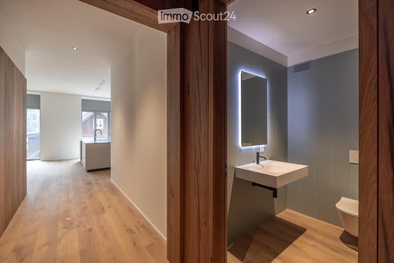 Modern bathroom with wooden flooring, a floating sink, a large illuminated mirror, and a toilet. The space is bright with recessed lighting and sleek, contemporary design.