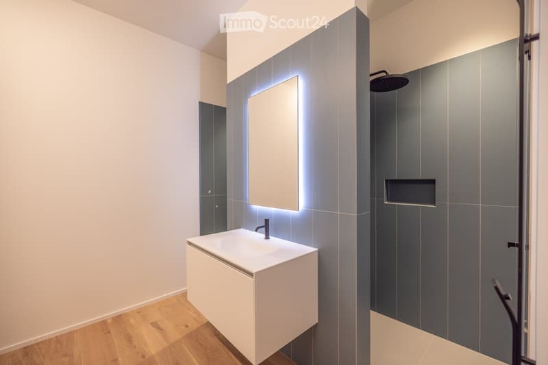 Modern bathroom with a white sink, dark grey tiled walls, a large mirror with built-in lighting, and a showerhead.