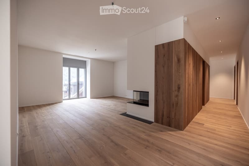 Empty room with light wooden flooring, white walls, recessed lighting, and a fireplace with a glass enclosure. A large window with a sliding door leads to a balcony.