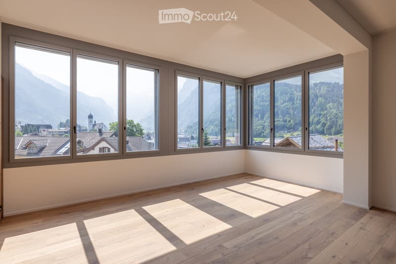 Empty room with wooden flooring and large windows offering a scenic mountain and village view.