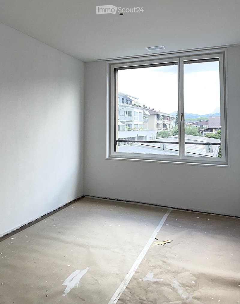 Empty room with bare walls and concrete floor, featuring a large window with a view of a building and mountains.