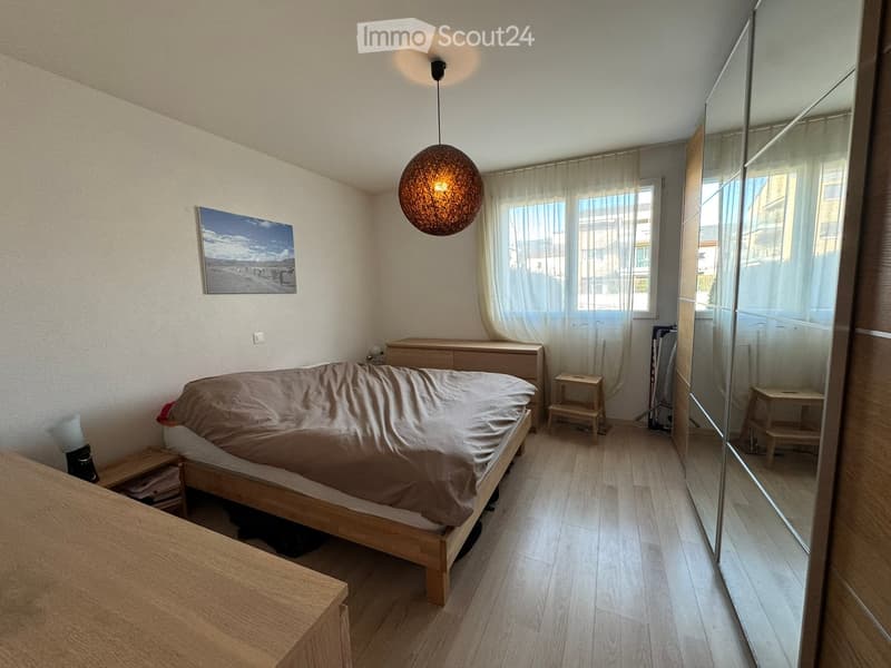 Modern bedroom with a wooden bed, nightstand, mirror, and pendant light. The room features light wooden flooring and large windows with sheer curtains.