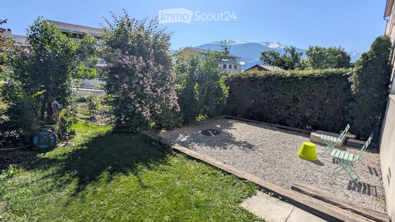 Garden with a gravel patio, green lawn, and a view of mountains in the background. Includes a folding chair and a yellow bucket on the patio.