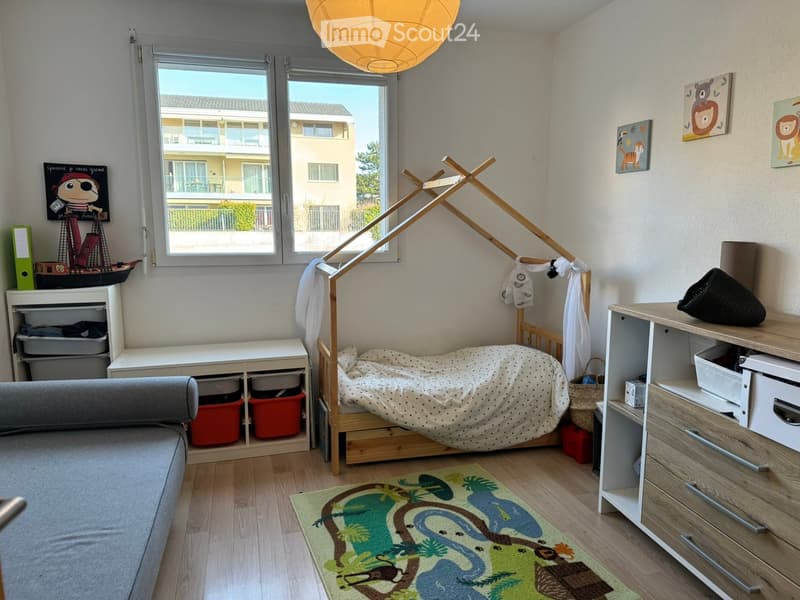 A well-lit bedroom with a wooden bed, a grey sofa, a nightstand, a storage unit, and a colorful rug with a dinosaur design. The room features large windows with a view of the outside and several decorative items including a toy ship and picture frames.
