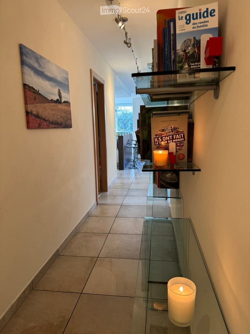 A modern hallway with tiled flooring, glass shelves holding books and candles, and a painting on the wall. A doorway leads to another room with visible furniture.