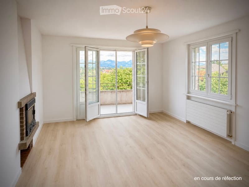 Empty room with light wooden flooring, white walls, large windows, and a fireplace. The room features a modern pendant light fixture and an open sliding door leading to a balcony with a scenic mountain view.