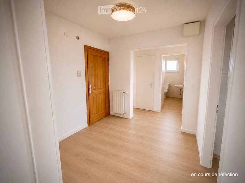 Empty hallway with light wooden flooring, white walls, and a wooden door leading to a bathroom visible at the end.