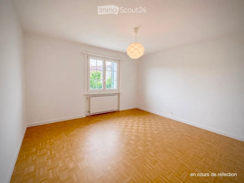 Empty room with light-colored walls, wooden herringbone flooring, a single window with white frame, radiator below the window, and a modern pendant light fixture hanging from the ceiling.