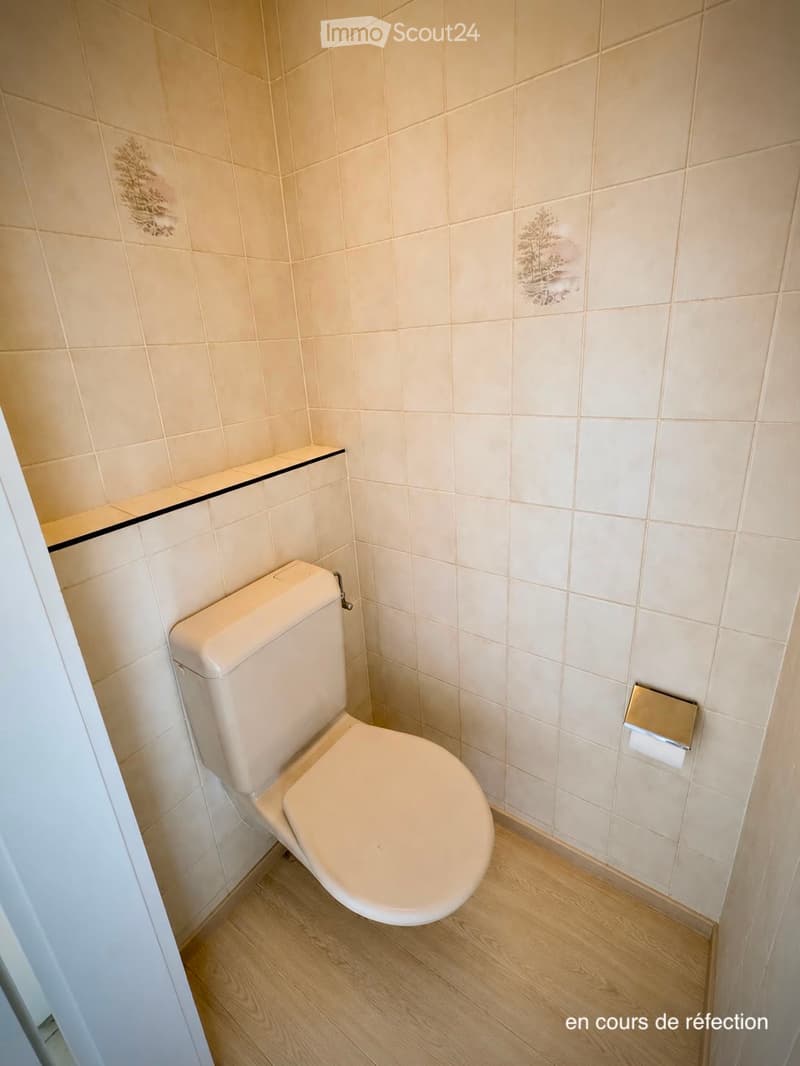 A small bathroom with beige tiled walls, a white toilet, and a wooden floor.