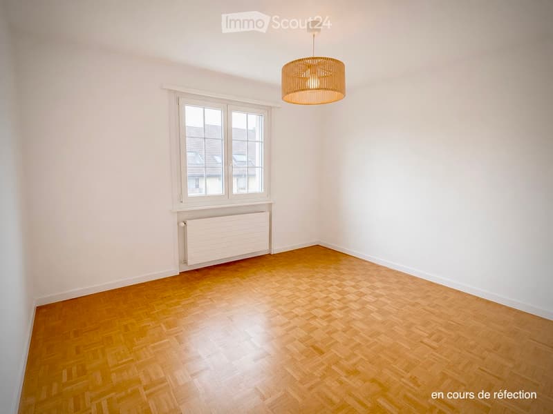 Empty room with wooden herringbone flooring, white walls, large window with white frame, radiator, and a hanging woven pendant light fixture.