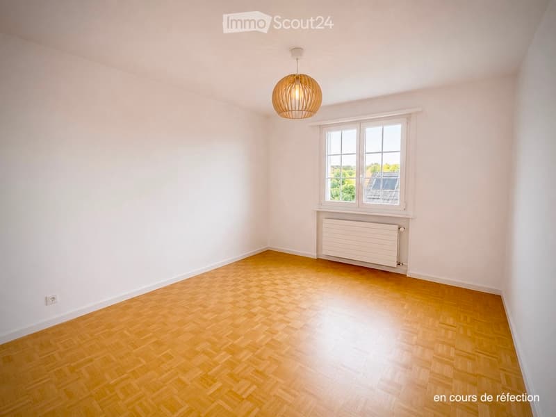 Empty room with light wooden herringbone flooring, white walls, a large window with white frame, a radiator below the window, and a modern pendant light fixture hanging from the ceiling.