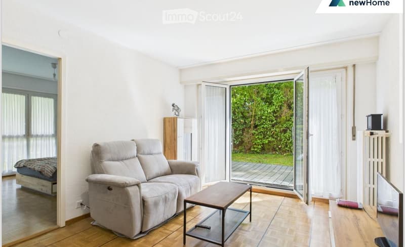 Living room with a grey sofa, coffee table, TV, radiator, and a view of greenery through large windows.
