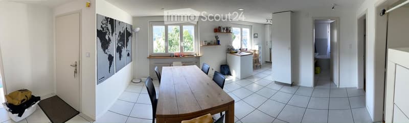 Bright, open-plan living and dining area with tiled flooring, large windows, a wooden dining table with chairs, a small kitchen island, shelves, and a world map on the wall.