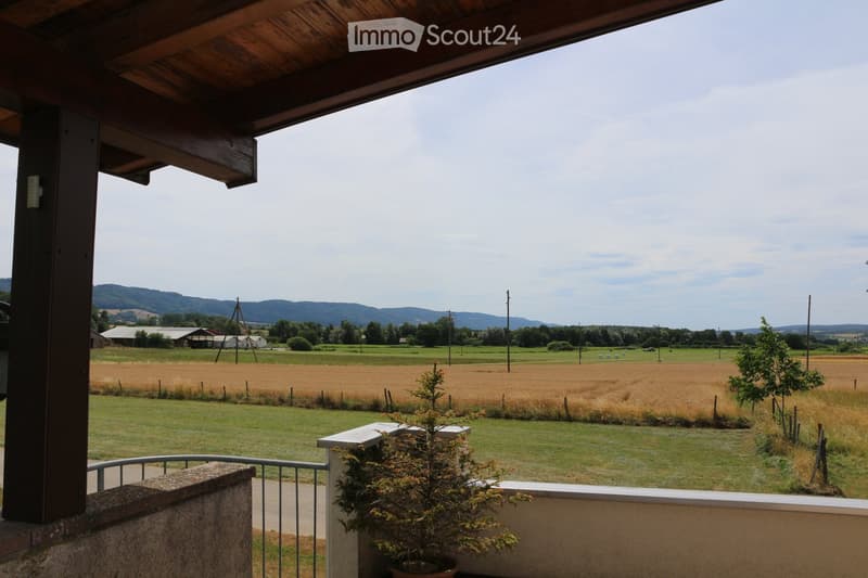 Balcony with wooden ceiling and metal railing, overlooking a rural landscape with fields, trees, and distant hills.