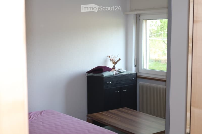 A small, well-lit bedroom with a bed covered in pink bedding, a wooden bench, a black nightstand with a vase and folded blankets, and a radiator beneath a window with a view of greenery.