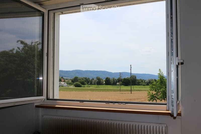 View from a window showing a rural landscape with fields, trees, and distant hills under a clear sky.