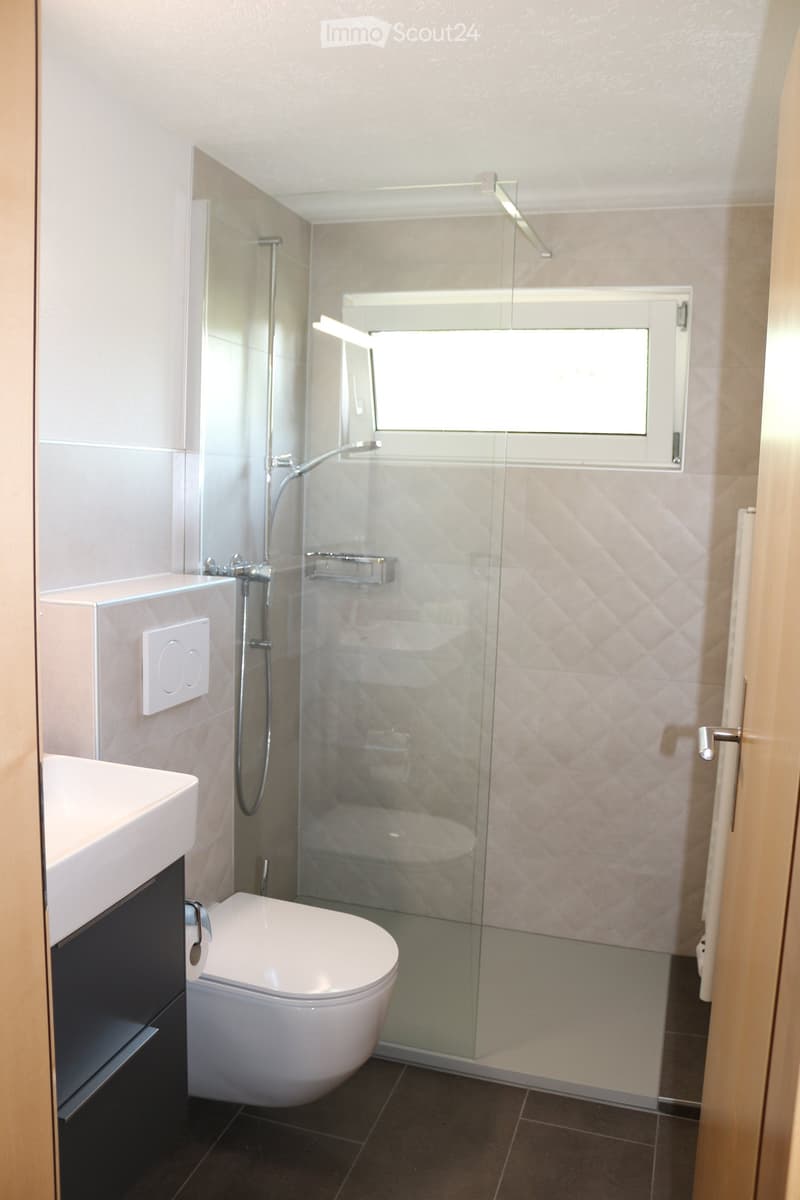Modern bathroom with a white toilet, sink, and glass-enclosed shower. Features a dark tiled floor and light-colored walls.