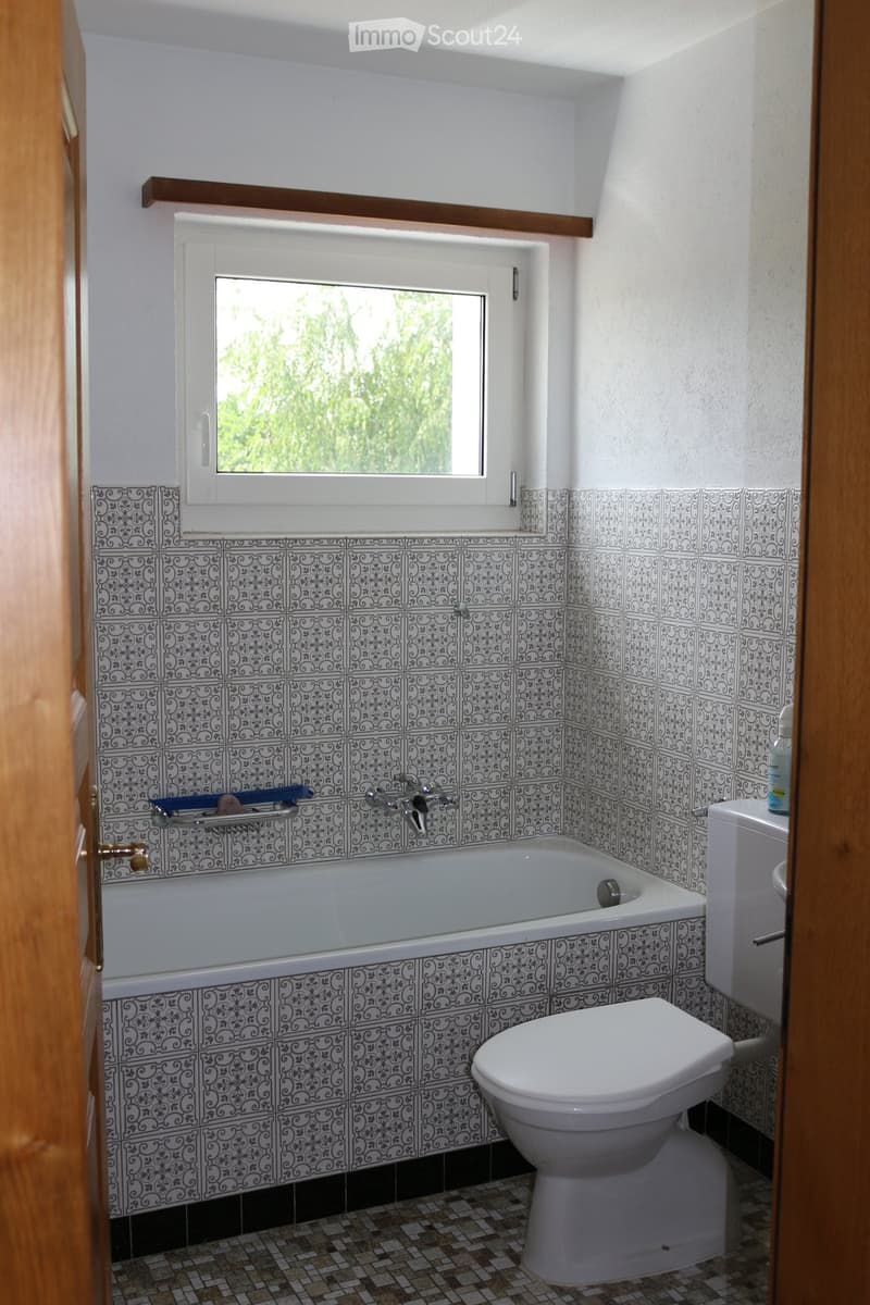 Bathroom with a bathtub, toilet, and ornate black and white patterned tiles. A window with a view of greenery is above the bathtub.