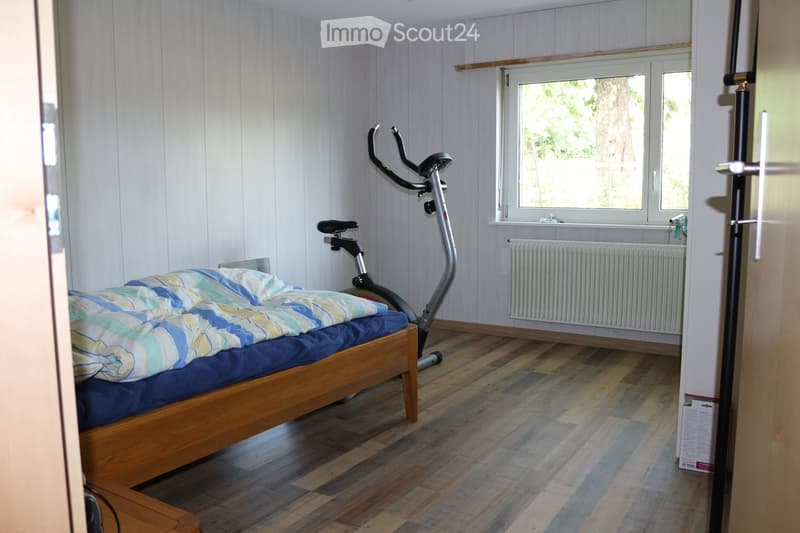 Cozy bedroom with wooden flooring, a single bed with a blue and green striped duvet, and an exercise bike positioned near the window. The room features light wooden panel walls and a radiator below the window.