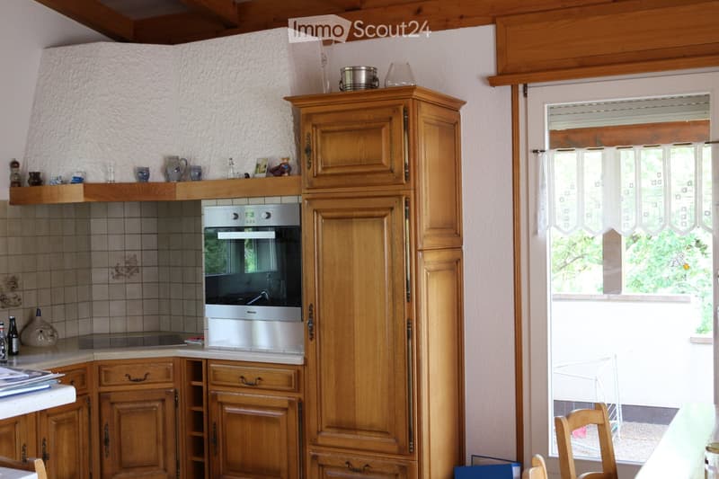 A cozy kitchen with wooden cabinets, a stainless steel oven, and a tiled backsplash. The countertop holds various items including cups, bottles, and a picture frame. A window with white curtains lets in natural light.