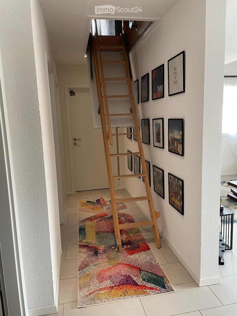 A narrow hallway with a colorful rug, a wooden easel, and several framed pictures on the wall. The hallway has white walls and tiled flooring.