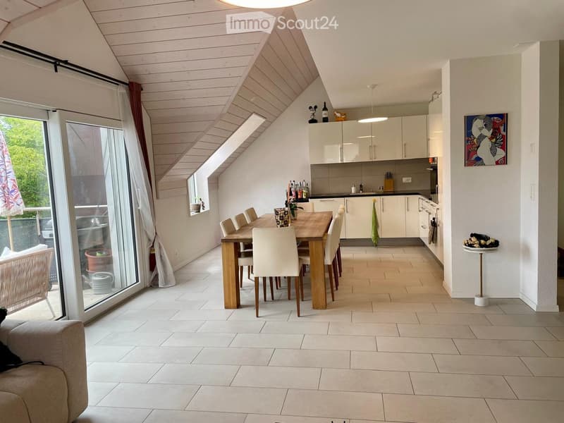 Open-plan living and dining area with a modern kitchen, dining table with chairs, and a sofa. The space features large windows with curtains, a painting on the wall, and a small stool with a decorative item.