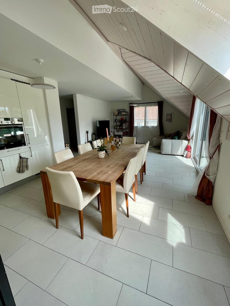 Bright, open dining area with a wooden dining table and white chairs, adjacent to a modern kitchen with white cabinets and an oven. A living area with a sofa and TV is visible in the background.