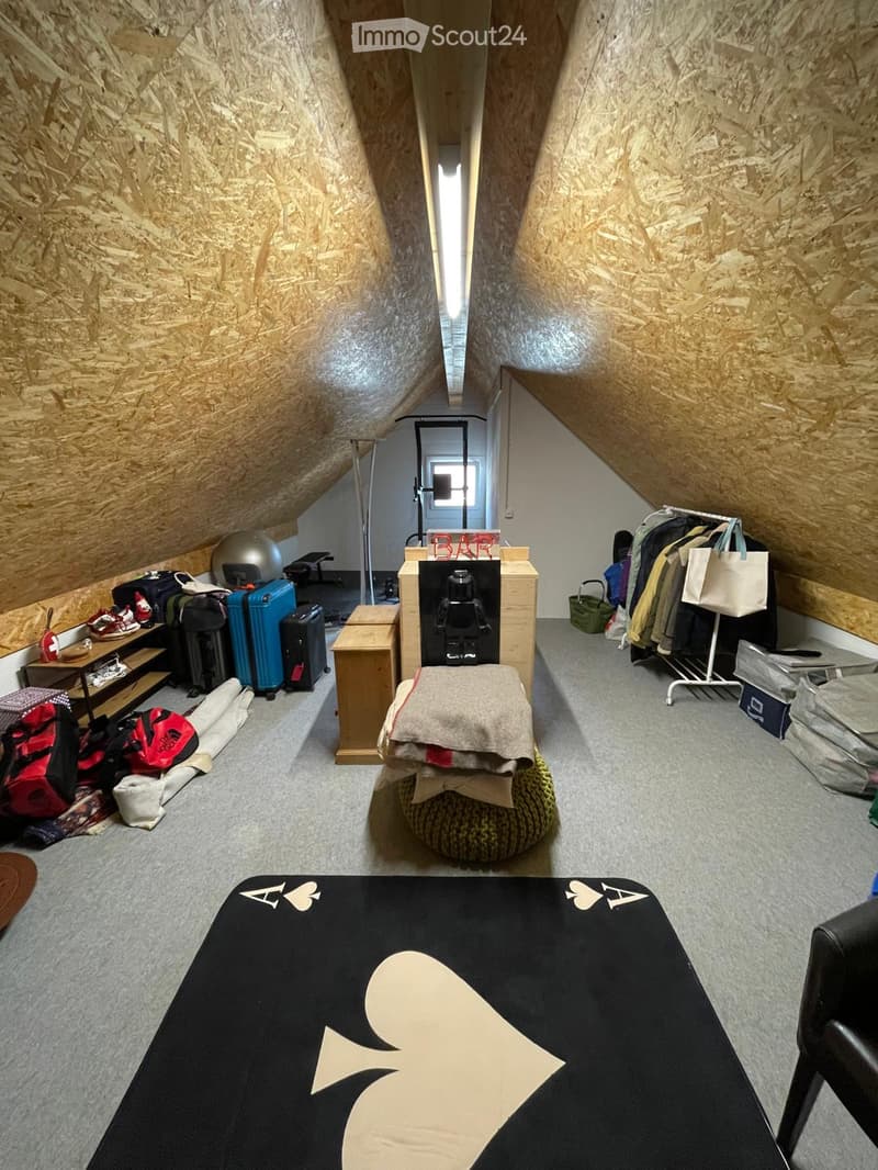 Attic room with wooden slanted ceilings, grey carpet, various luggage and bags, a bar area, a round stool with a blanket, and a clothing rack with bags.