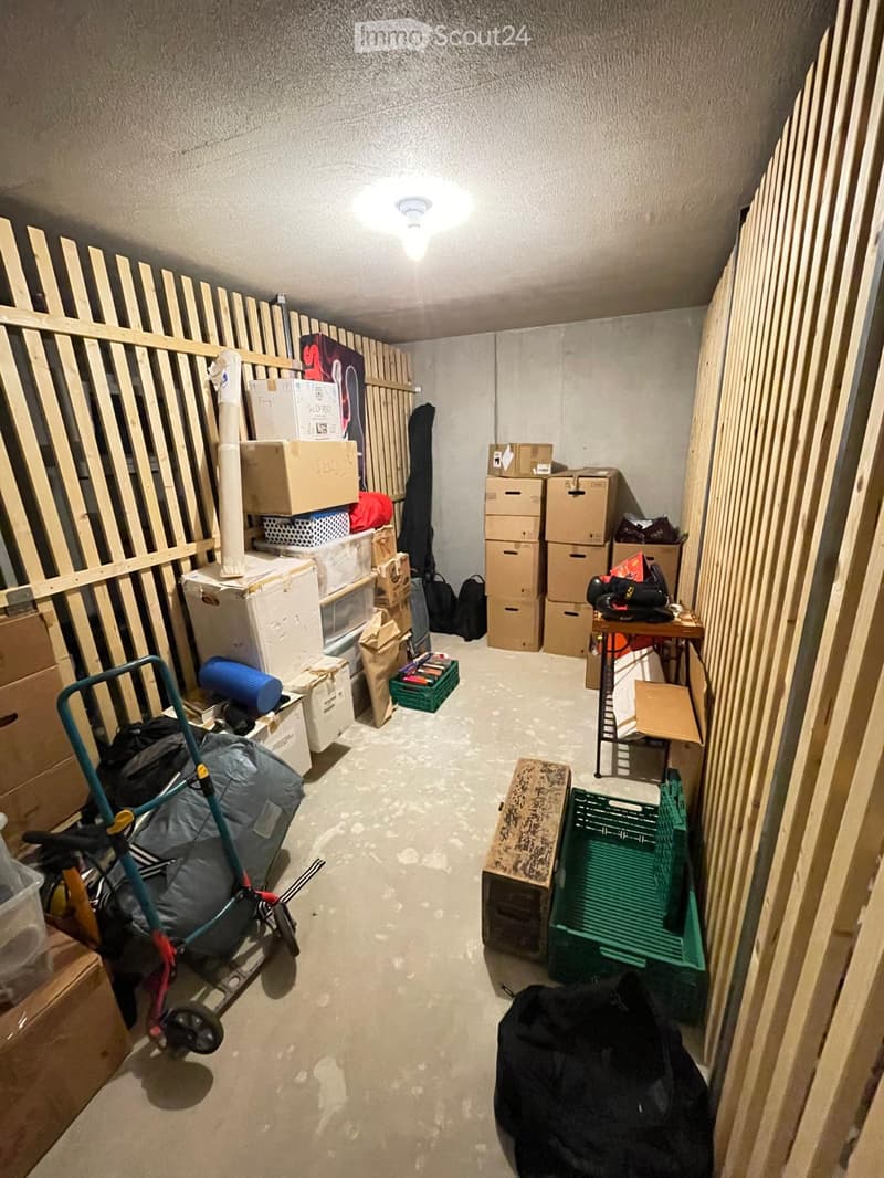 A narrow, concrete-walled corridor with a ceiling light, lined with stacked cardboard boxes, plastic containers, and miscellaneous items including a rolling cart, bags, and a table with items on it.
