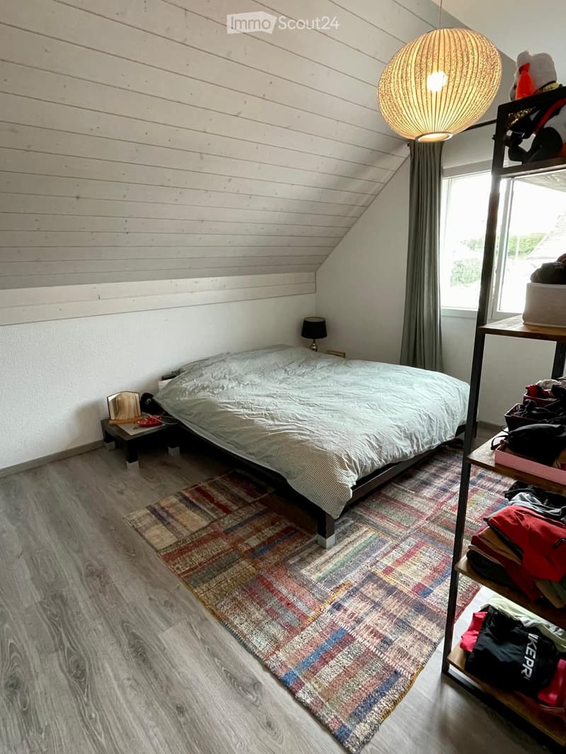 Cozy attic bedroom with wooden walls, slanted ceiling, and large window. Features a bed with a patterned duvet, a nightstand with a lamp, a colorful rug, and a shelf with folded clothes and bags.