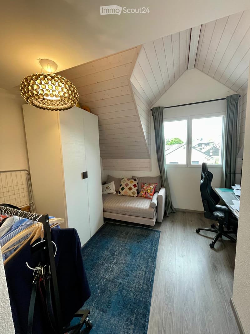 A cozy, multi-functional room with a slanted wooden ceiling, a small sofa with decorative pillows, a desk with a chair, a wardrobe, and a hanging rack with clothes.
